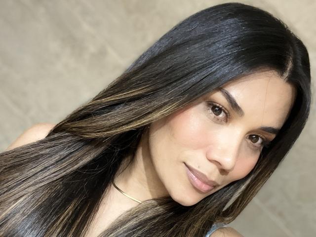Sheyleeya has long, straight black hair with subtle brown highlights, brown eyes, and a slight smile. She's wearing a thin gold necklace. Her skin is light brown.