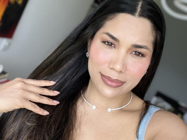Sheyleeya has long black hair, brown eyes, pink blush, and dark pink lipstick. She's wearing a silver necklace with small diamonds, and a blue sleeveless top.