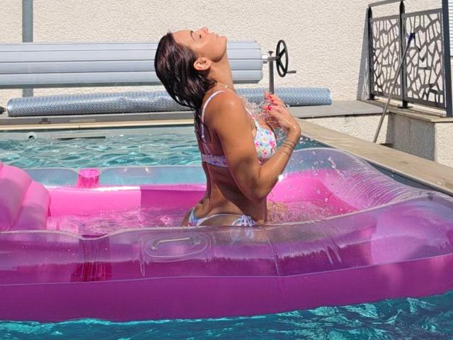 JuneLaFrenchie, wet and tan, kneels on a pink inflatable pool toy, head tilted back, eyes closed, hands on chest. Water splashes around, pool edge, sunlit, seductive pose.