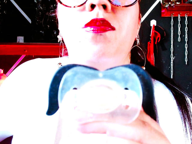 LexyDom, con lápiz labial rojo, sostiene un juguete sexual azul con forma de cerdo en primer plano. Lleva gafas y grandes aros, y tiene el cabello negro. Está muy bien iluminada..