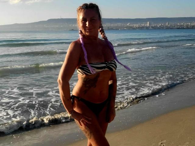 ChaudeEvely stands on a beach, wearing a zebra-striped bikini, purple braids, and black bottoms, with a tattoo on her right side, and her tan, toned body is visible.