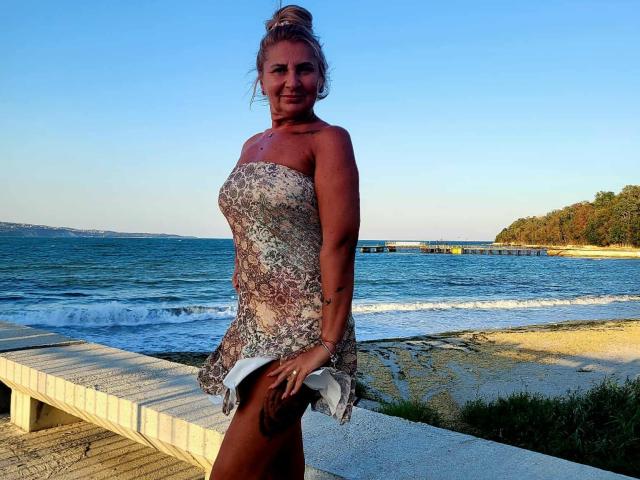 ChaudeEvely with tan skin, blonde bun, wearing a strapless, floral dress, standing on a wooden bench by the beach, holding a white towel, showing her small tits.