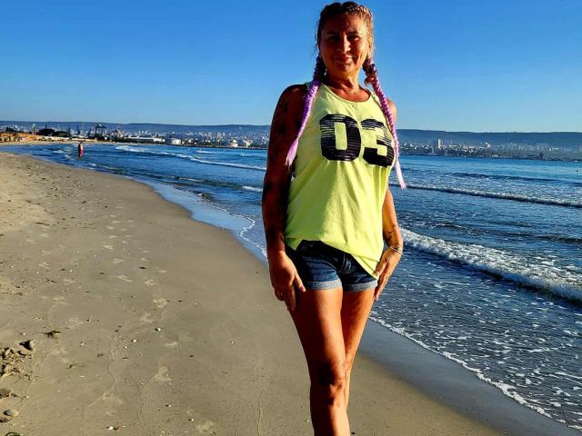 ChaudeEvely with blonde braids, yellow tank top, and blue shorts, standing on a sandy beach, wearing a number 03 jersey, and showing her tanned, tattooed arms.