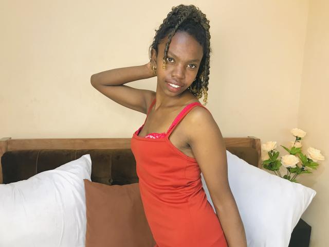ElianeJolie with dark skin, curly black hair, and red dress, standing, arm behind head, smiling, small tits, brown eyes, white pillows, brown cushion, white roses in background.