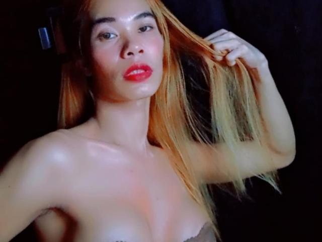AsianStarTS with shiny skin, red lips, and blonde hair, holding her hair up, showing her tits, against a dark background. Her boobs are small and she's topless.