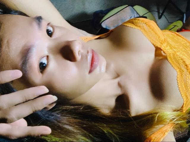 AsianStarTS lying down, wearing an orange lace bra, eyes looking up, hand touching her head, medium tits, long brown hair, light skin, subtle makeup, slight smile, relaxed.