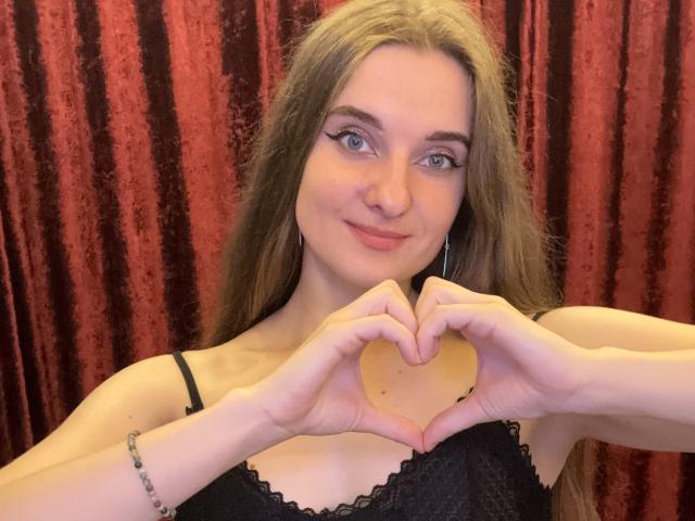 KariMillie with long brown hair, making a heart shape with her hands, wearing a black lace top, smiling, and showing small tits against a red curtain.