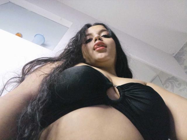KonyBella, com cabelos longos e pretos, olhando para a câmera, vestindo um sutiã preto com um recorte em formato de chave, mostrando seus seios grandes e arredondados. Ela tem um leve semblante de beicinho..