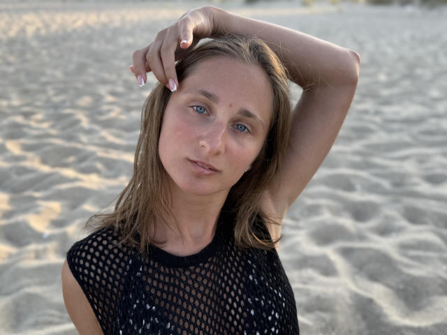 MichelleHotX69 with blue eyes, wearing a black fishnet top, has her arm raised, showing her small tits and a slightly flushed face, on a sandy beach.