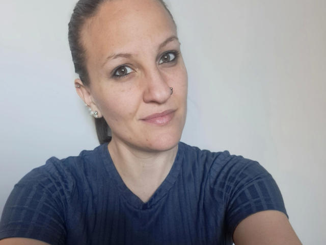 Swissmiss88-hot, a woman with brown hair in a ponytail, wearing a navy blue V-neck shirt, has a nose ring, small earrings, and a subtle smile. Her tits are average.