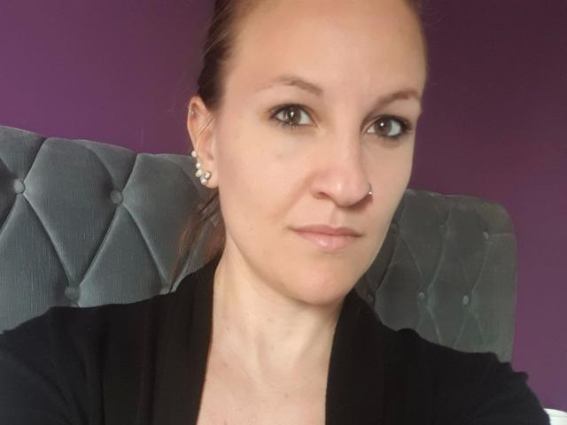 Swissmiss88-hot has a nose piercing, small tits, and is wearing a black shirt with pearl earrings, against a purple background and grey tufted headboard.