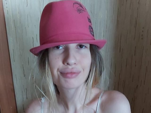 ElzaKomx with blonde hair, pink hat, and slight smile, wearing a white top, has small tits and light skin, standing in front of a wooden wall.