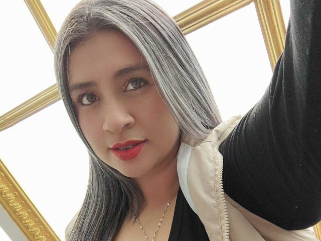 MelindaBoobs with gray and white hair, red lipstick, black and white jacket, and a nose ring, taking a selfie, showing a slight smile and looking at the camera.