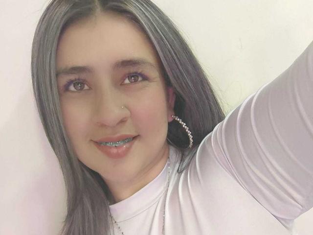 MelindaBoobs with long gray hair, white shirt, nose ring, and pink hoop earrings, smiling with glittery blue lips, taking a selfie.