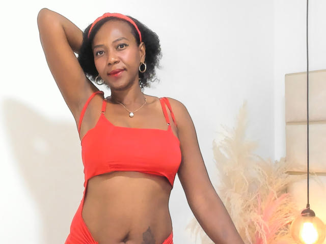 AfroHeat mit dunkler Haut, rotem Stirnband, rotem BH und roter Unterwäsche, Arm erhoben, zeigt kleine Brüste und flachen Bauch, Ohrringe, Glühbirne und eine buschige Pflanze im Hintergrund..