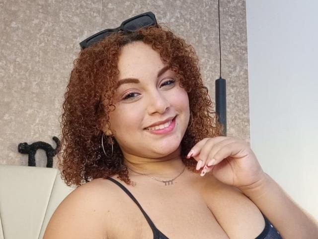 YeraCarey with curly brown hair, smiling, showing her tits in a black strap top, wearing hoop earrings, and a necklace with "MM" in the center.
