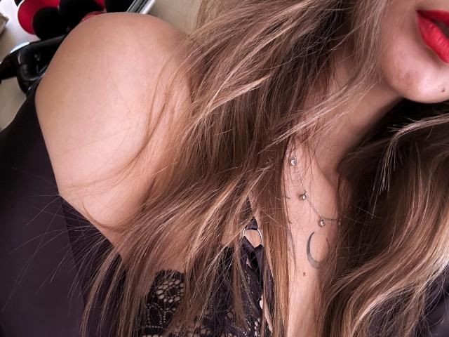 Karish's bare shoulder, long blonde hair, red lips, black lace top, and a crescent moon necklace. Boobs partially visible, soft light. Slightly messy hair, sensual vibe.