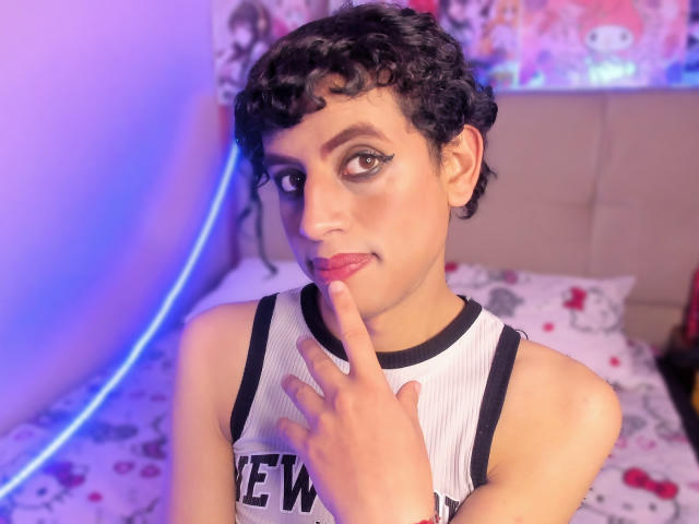 EimyFlorian with curly black hair, wearing a white tank top, has finger near pink lips, brown eyes with winged eyeliner, on a bed with colorful patterned sheets.