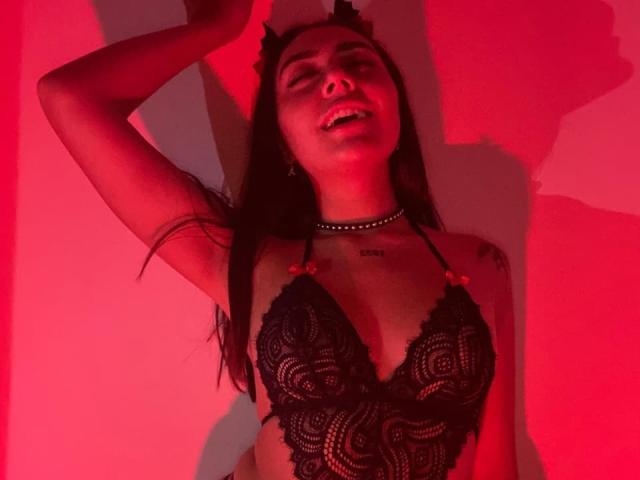 AlannaJonson with long brown hair, wearing a black lace bra, choker, and devil horns, smiling, eyes closed, under red lighting, arm raised. Boobs visible through lace.