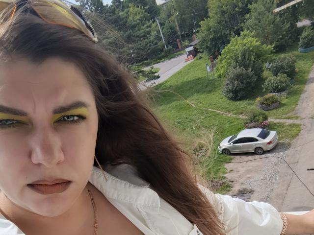 Almazy's face with yellow eyeliner, brown hair, white shirt, and brown lipstick, looking annoyed. White car parked on gravel, green grass, and trees in background. Slightly blurry.