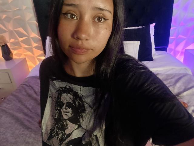 Karollteyes with long black hair, brown eyes, and a cute face, wearing a black shirt with a rock star print, sitting on a bed, looking at the camera.