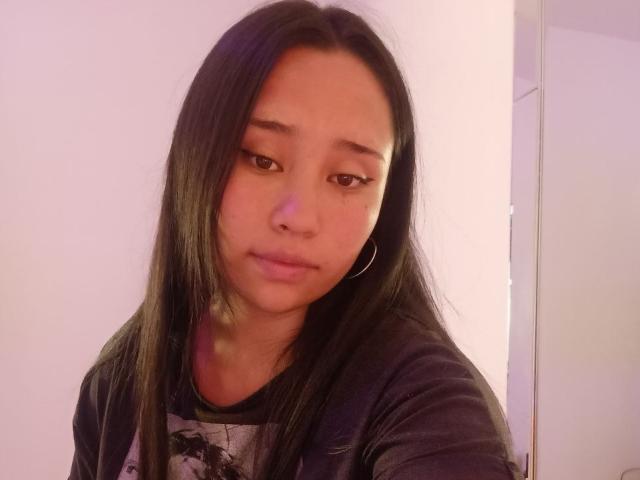 Karollteyes with long black hair, wearing a dark shirt, has small hoop earrings, brown eyes, and a slightly pouty expression, looking off to the side.