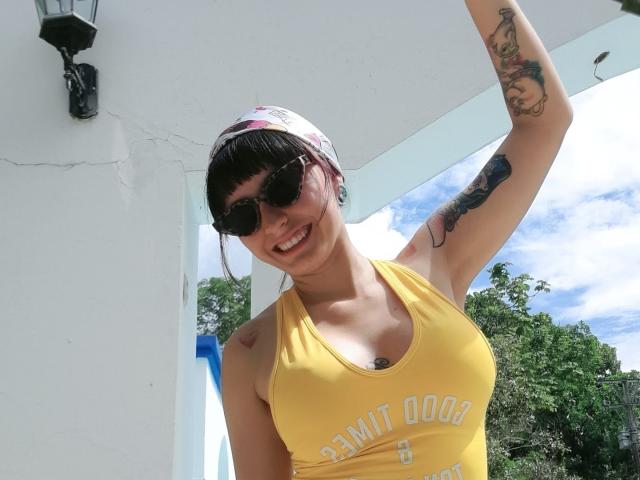 SaraleePoison, wearing a yellow tank top, black sunglasses, and a white cap, smiling with her arm raised, showing tattoos on her arm and side, sunny day, cute.
