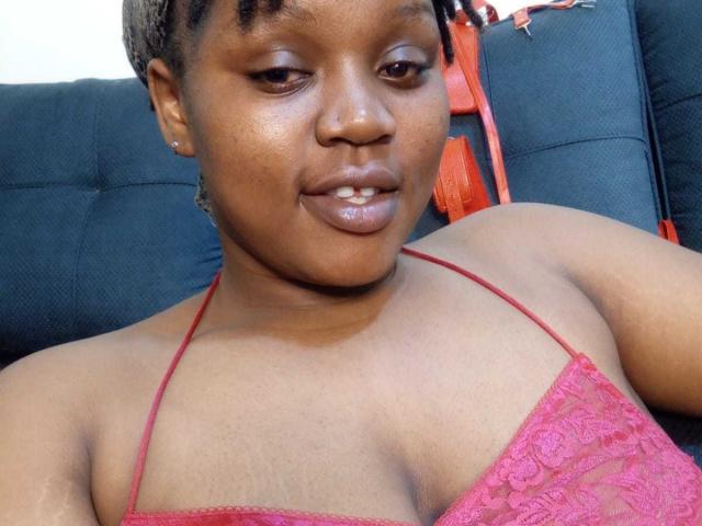 InfinityCouple with short, braided hair wearing a pink lace halter top, showing small tits and a subtle smile, sitting on a black couch. Her skin is dark brown.
