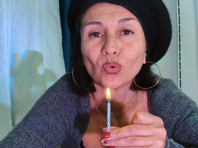 LatinLola pokes out her lips, holding a lit candle with a blue and white striped pattern, wearing a black hat, hoop earrings, and a gray cardigan.