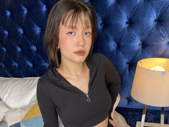 AraknyRoz with medium-length brown hair, black zip-up top, small tits, fair skin, pouty lips, blue tufted wall, white pillow, beige lamp, and necklace, looking at camera.