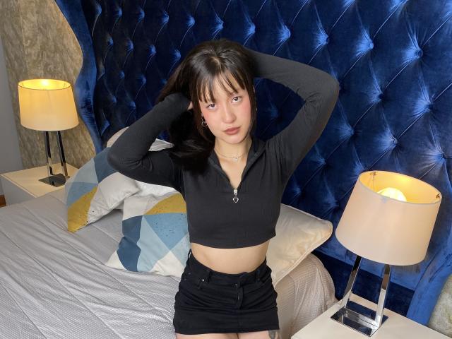 AraknyRoz with black hair, wearing a black crop top and black shorts, sitting on a bed with hands in hair, blue tufted headboard, and two lit lamps.