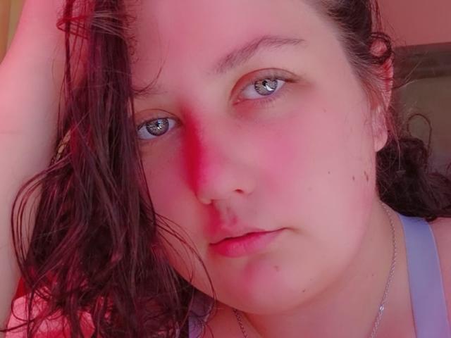FranRose's face with wet brown hair, blue eyes, pink blush, slight frown, wearing a gray tank top, and a silver necklace, under red lighting.