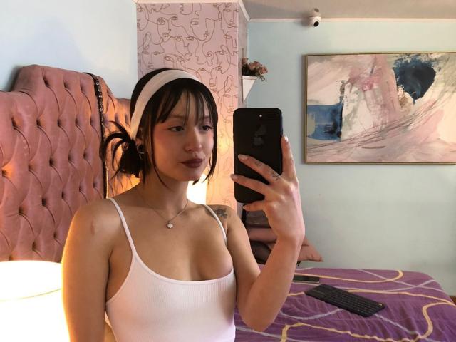 AraknyRoz with small tits, wearing a white tank top and white headband, taking a selfie in a bedroom with pink tufted headboard and purple bedspread. Holding a black phone.