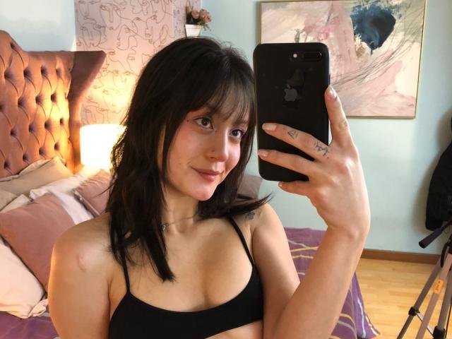 AraknyRoz with black bra, holding iPhone, smiling, showing small tits, tattoo on ring finger, dark hair, light skin, in bedroom with pink tufted headboard, purple bedspread.