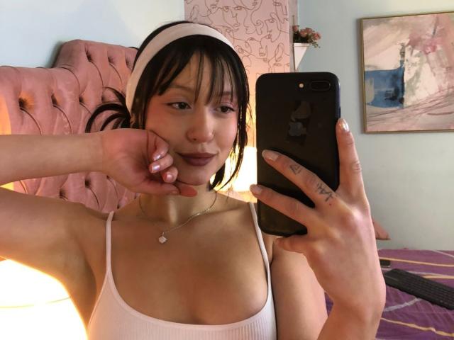 AraknyRoz with black hair, white headband, and pink lipstick, taking a selfie in a mirror. She's wearing a white tank top, showing her small tits, and holding a black iPhone.