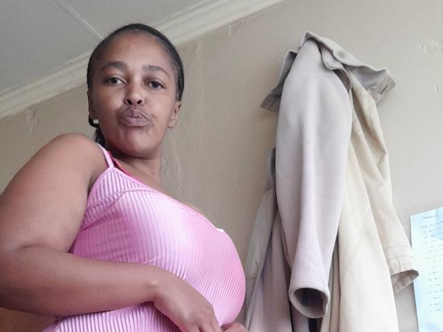 CassCecilia with dark brown skin, wearing a pink ribbed tank top, showing her boobs, puffy lips in a kissy face, beige jacket hanging behind her.