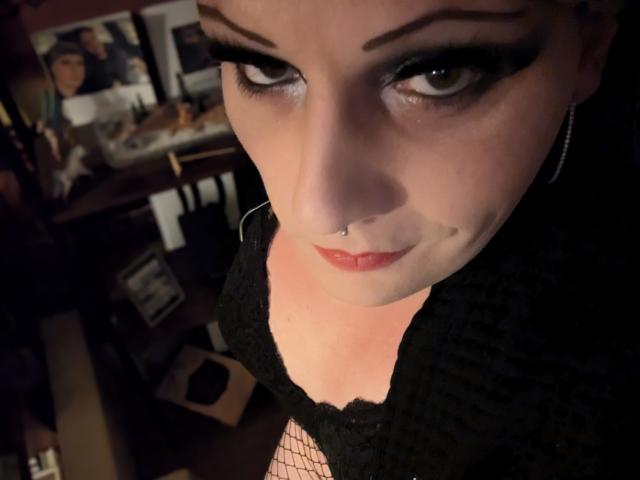JennaJoy69 with dark eyeliner, red lipstick, black lace top, nose ring, and fishnet sleeves, looking up with a slight smile, showing her boobs. Dimly lit room in background.