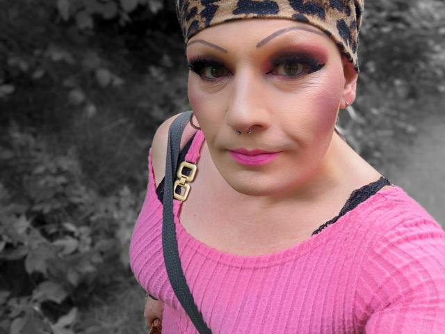 JennaJoy69 has pink lipstick, thick black eyeliner, leopard print headscarf, nose ring, pink ribbed shirt, black lace strap, and a confident smile, with a gray, leafy background.