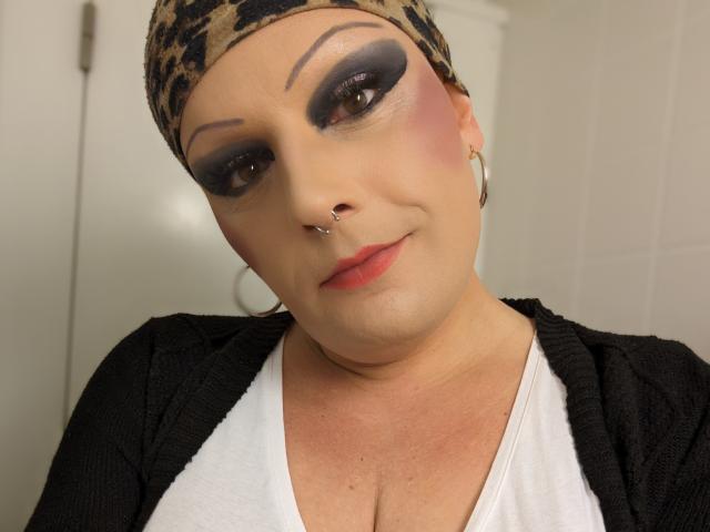 JennaJoy69 with heavy black eyeliner, purple eyeshadow, pink blush, nose piercing, hoop earrings, leopard print headscarf, black cardigan, white top, and red lipstick, smiling softly.