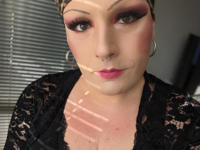 JennaJoy69 with a nose piercing, pink lipstick, black lace top, hoop earrings, and dramatic eye makeup. Sunlight stripes her face. She's a chubby, white, sexy, cute, attractive woman.