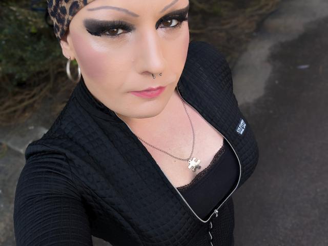 JennaJoy69 with leopard-print headscarf, black eyeliner, pink blush, nose ring, black zip-up, cleavage showing black top, silver necklace, and hoop earrings, looking up at camera.