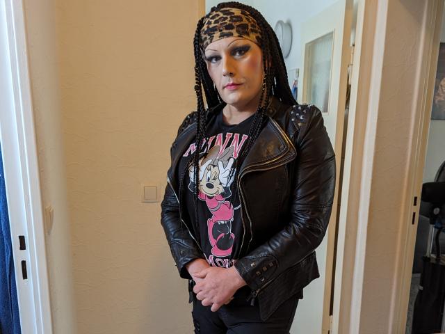JennaJoy69 stands with hands clasped, wearing a black leather jacket, "Plum Puff" shirt, black pants, and leopard print headscarf, with braided hair and subtle makeup, looking serious.