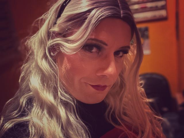 JennaJoy69 with wavy, silver-blonde hair, dark eyeliner, and red lipstick, smiling slightly. She's wearing a black headband and dark top, in a warmly-lit indoor setting.