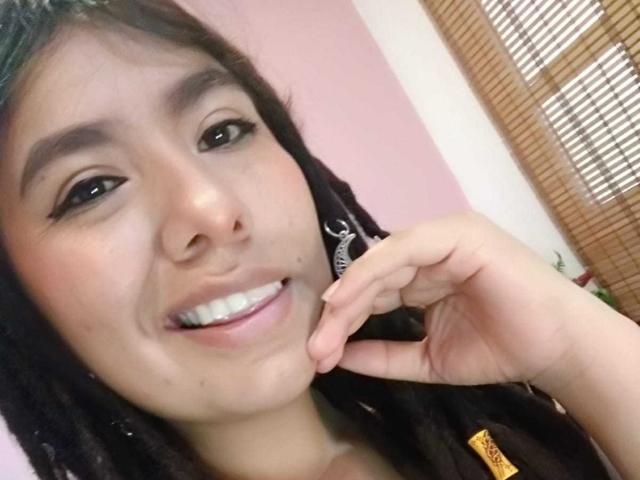 BeckyHuston has dark brown hair, black eyeliner, and a nose ring. She's smiling with her finger near her cheek, showing white teeth and a small beauty mark.