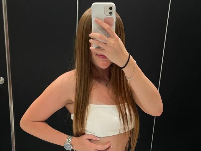 ChicSparkle mit langen braunen Haaren, trägt ein weißes Top und macht ein Selfie mit ihrem Handy, wobei ihre Brüste und ihre Hand auf ihrem Bauch zu sehen sind. Sie trägt ein Armband..