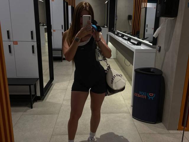 ChicSparkle mit langen braunen Haaren, trägt ein schwarzes Sport-Top und Shorts, hält eine Kamera, zeigt ihr Gesäß und ihre Brüste in einem Spiegel-Selfie, eine weiße Tasche mit der Aufschrift "FUTBOL" hängt ihr über der Schulter..