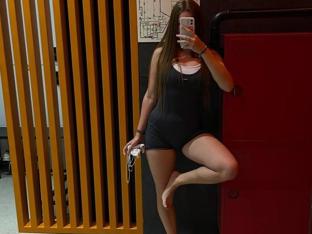 ChicSparkle mit langen braunen Haaren, in einem schwarzen Einteiler, Schlüssel in der Hand, stehend, ein Bein angehoben, Spiegelbild, roter Spind, Holzlatten, Telefon in der Hand, zeigt ihre Brüste und ihren Hintern..