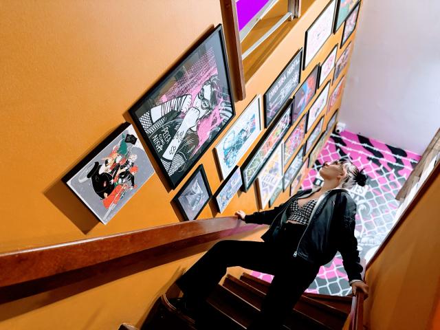 SaraleePoison leaning against a wooden staircase, wearing a black jacket and black pants, looking at a wall of colorful framed art. Her ass is slightly raised.