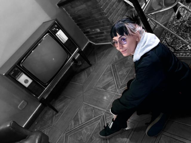 SaraleePoison, crouching, wearing glasses, black jacket, white hoodie, black sneakers, with short black-and-white hair, in a room with a vintage TV and patterned floor.