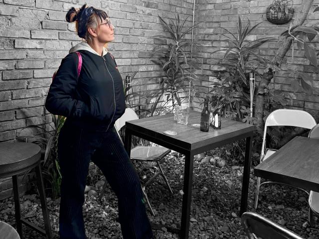 SaraleePoison stands in a gray-bricked outdoor area, wearing a black hoodie and pinstriped pants, hands in pockets, with blue hair in a bun, looking up.